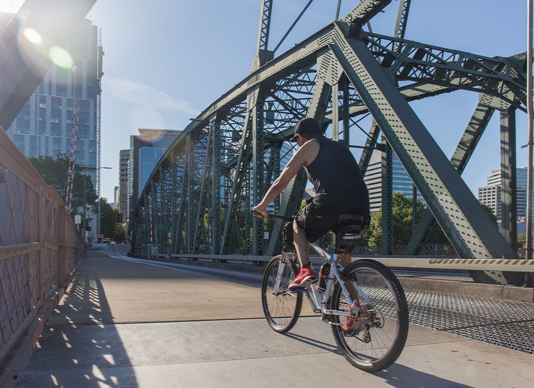Oregon City, OR - Man Riding Bicycle Across Bridge Against Portland Cityscape on a Sunny Afternoon